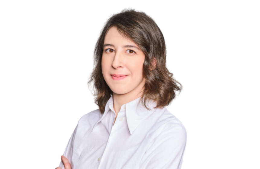  The photo shows content writer at Primotly. A contented-looking woman with medium-length brown hair stands with her arms crossed, smiling gently at the camera. She is wearing a crisp white shirt, suggesting a professional setting. Her posture and expression convey confidence and approachability. The background is plain white, highlighting her as the subject.