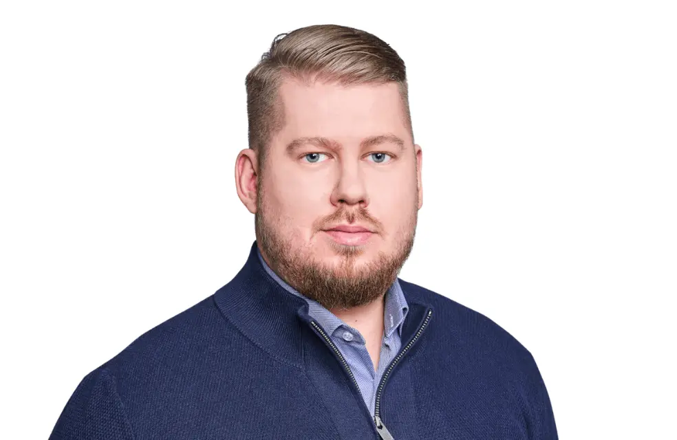 The photo of the article's author - CTO at Primotly.  The photo shows a self-assured man with neatly styled short hair and a trimmed beard gazes directly at the camera. He is wearing a sharp, buttoned-up blue shirt layered with a dark blue zippered sweater, presenting a polished and professional appearance. The soft white backdrop ensures he is the focal point of the image.